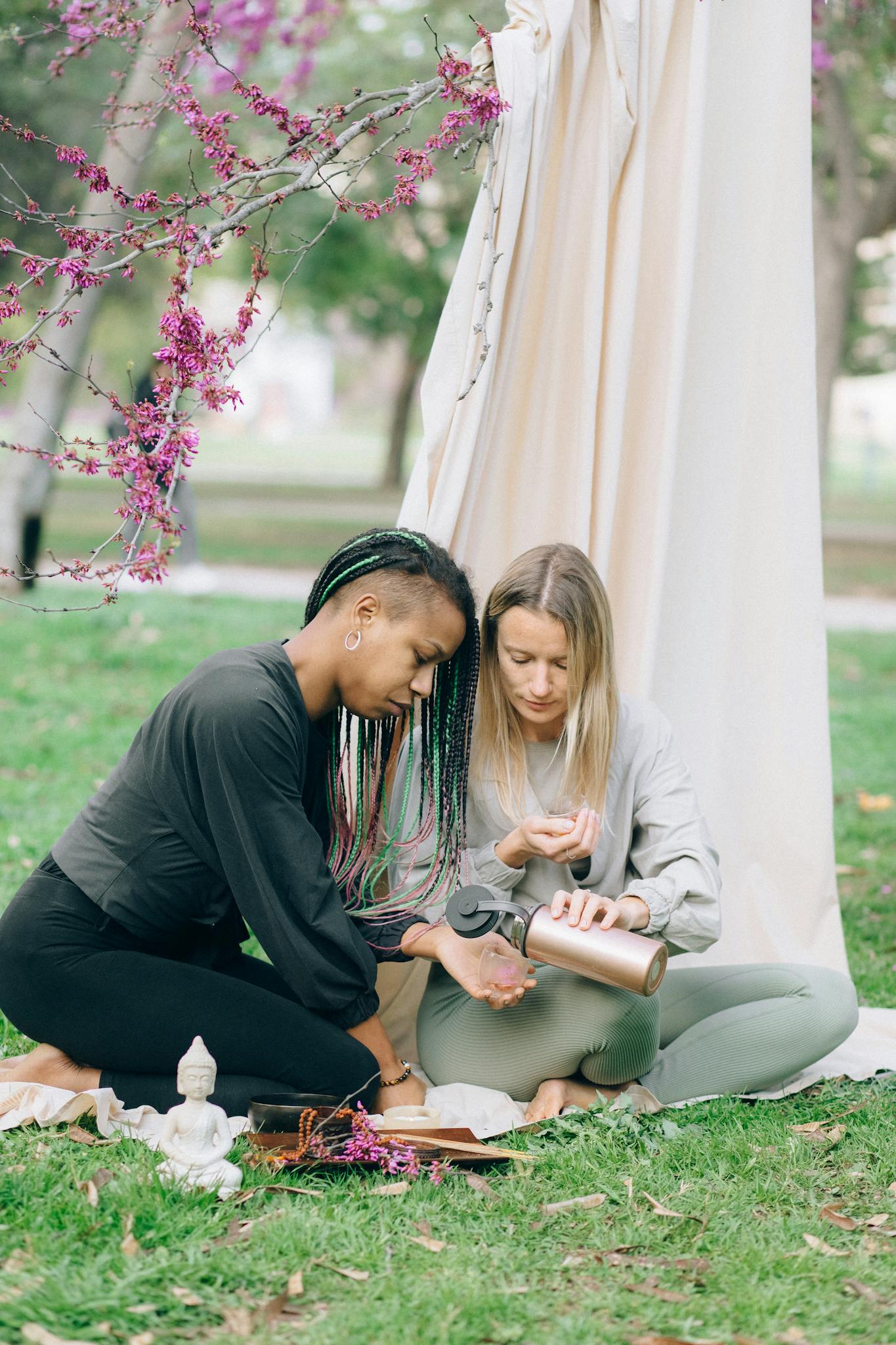 Two women relax on a peaceful outdoor picnic surrounded by nature, enjoying a warm beverage.